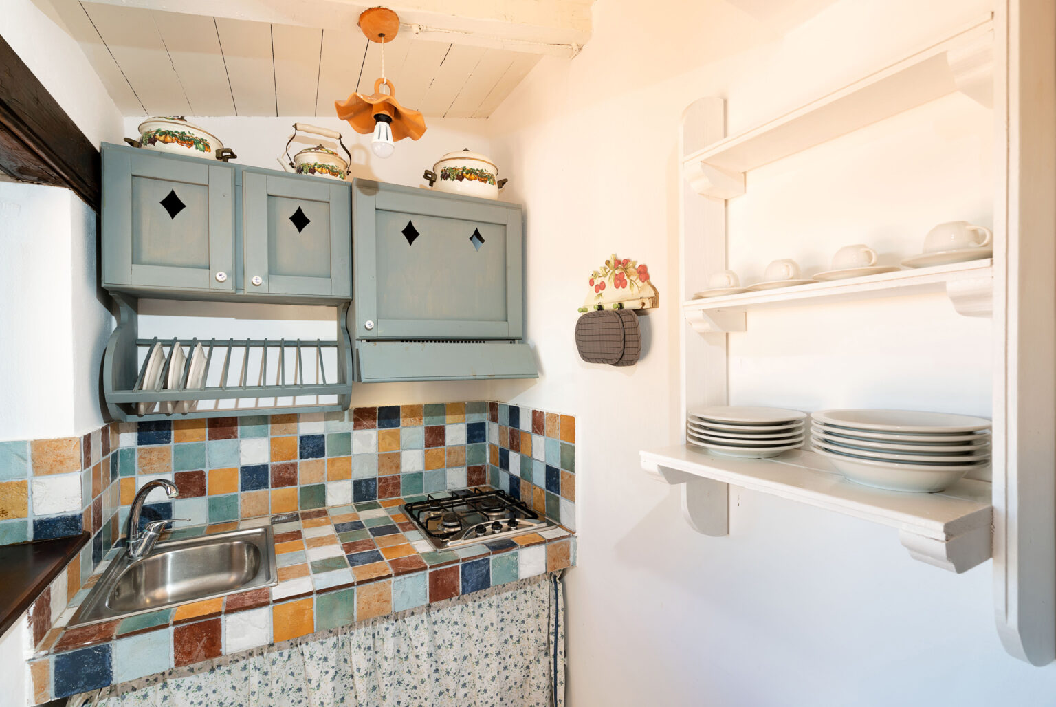 Cozy kitchen featuring blue cabinetry, a colorful tiled backsplash, and open shelving with plates, designed for efficient space utilization and a welcoming atmosphere. | Only on SopranoVillas