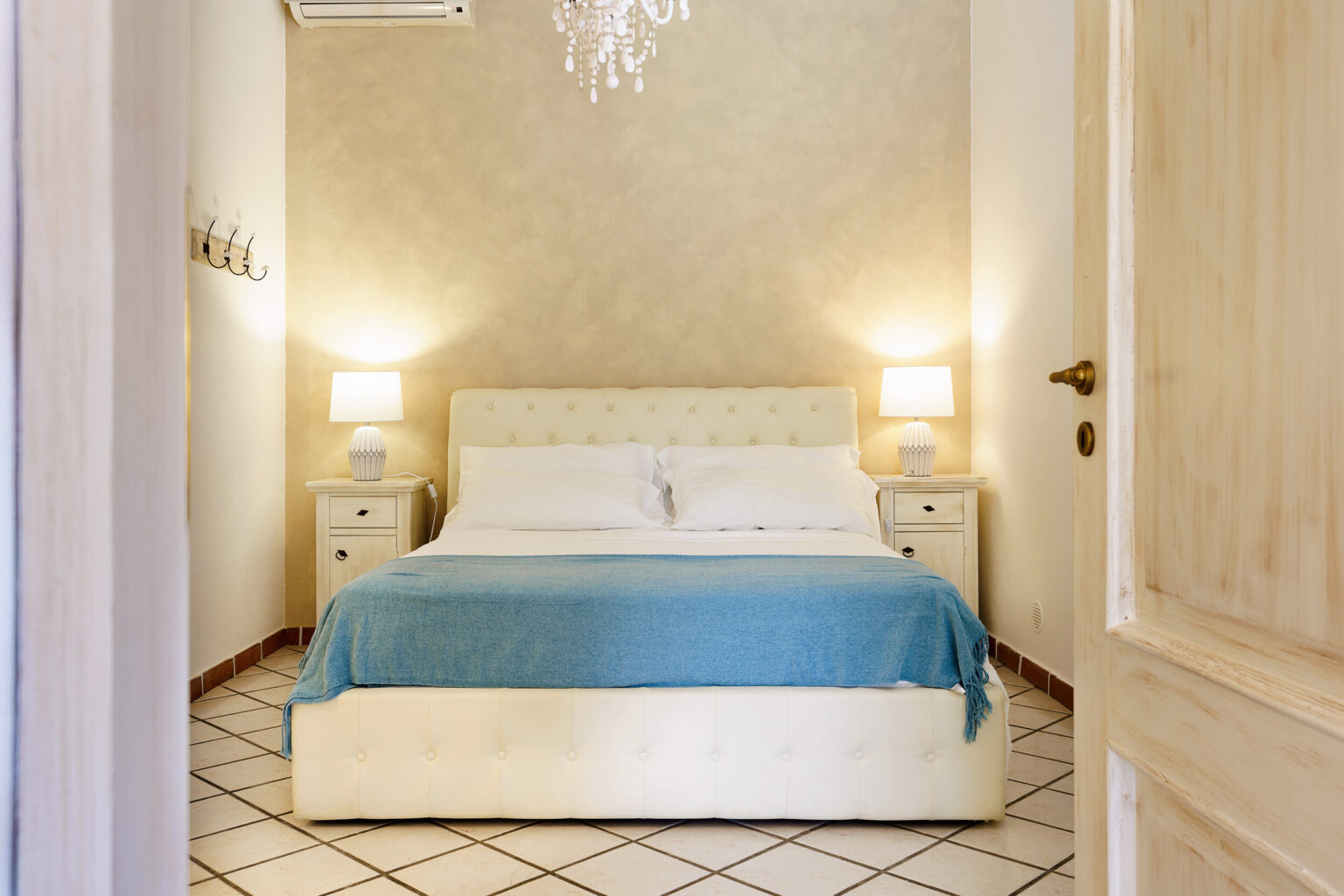 Cozy bedroom featuring a queen-sized bed with a light blue throw, bedside tables with lamps, and a chandelier, set against a warm, neutral wall. The room is illuminated and inviting, perfect for relaxation. | Only on SopranoVillas