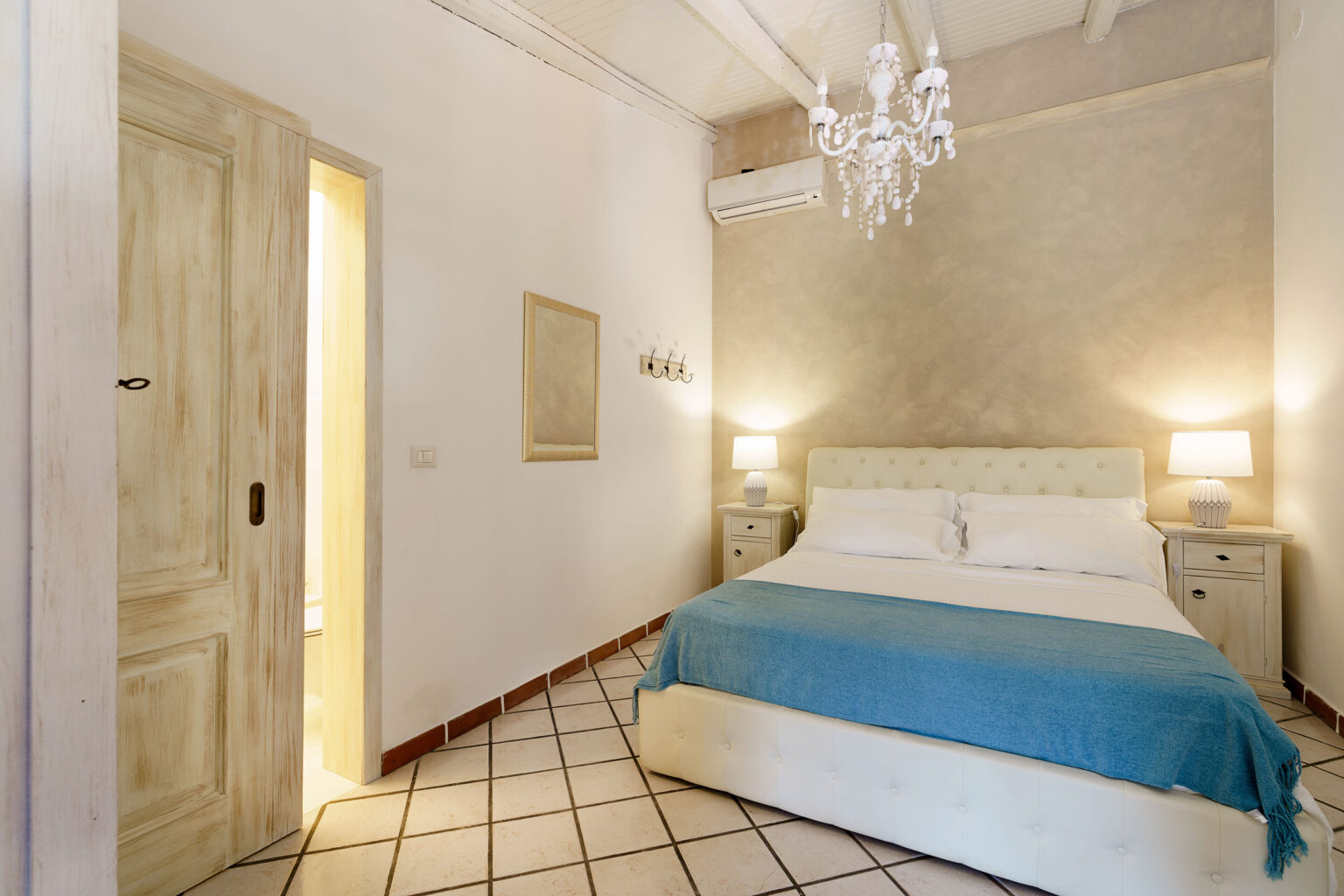 Cozy bedroom featuring a comfortable queen-sized bed with a light blue throw, elegant bedside tables, a chandelier, and a softly lit ambiance, ideal for relaxation. | Only on SopranoVillas