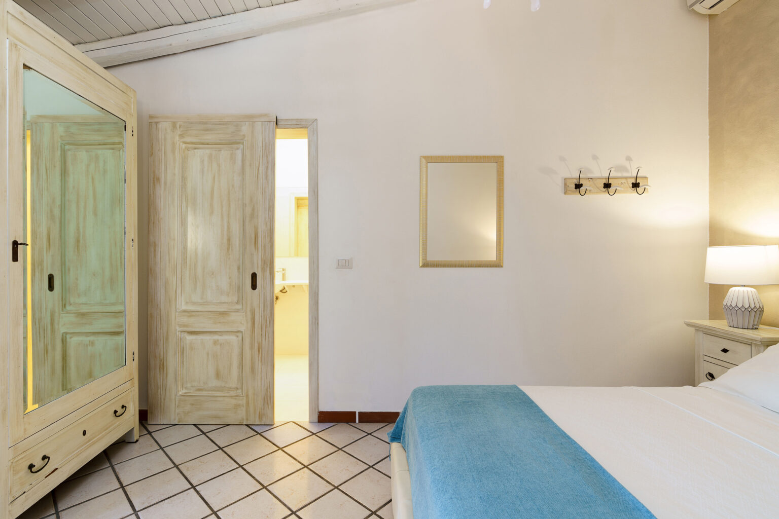 Cozy bedroom featuring a light-colored wardrobe, a bed with a blue throw, and a mirror on the wall, with an adjoining bathroom doorway visible. | Only on SopranoVillas