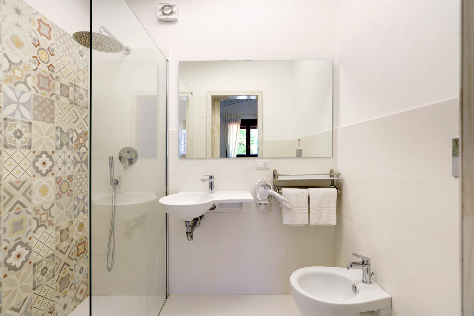 Modern bathroom interior featuring a glass shower, wall-mounted sink, bidet, and decorative tiled wall. The space is well-lit with a large mirror and minimalistic design, emphasizing cleanliness and functionality. | Only on SopranoVillas