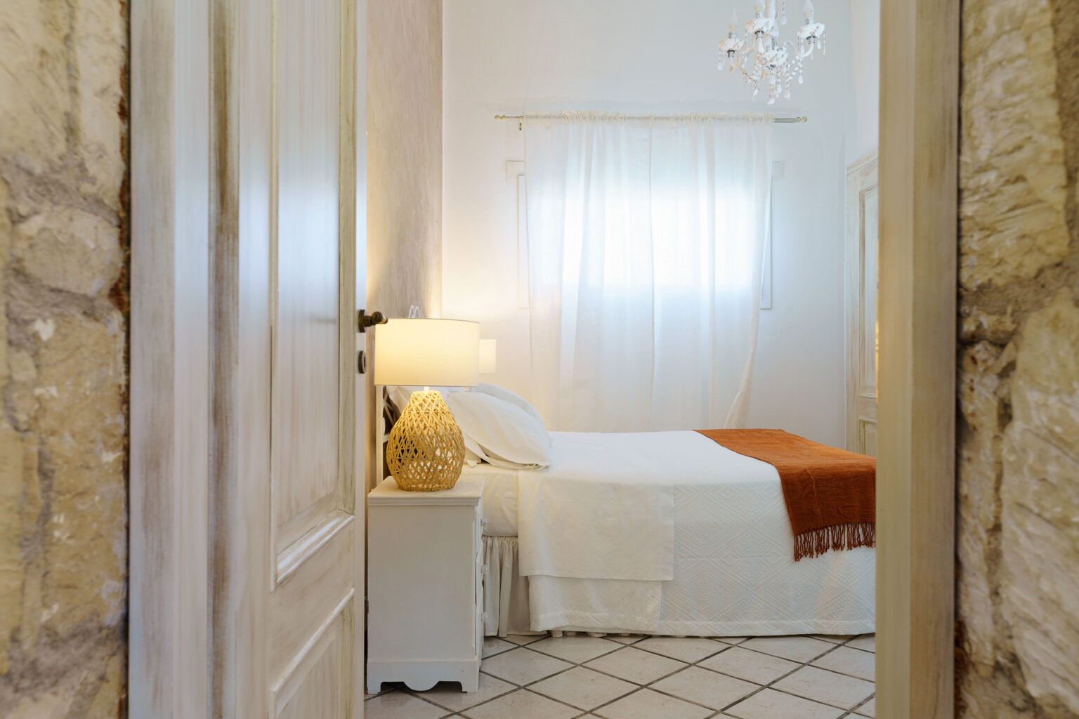 Cozy bedroom with a white bedspread and orange accent blanket, featuring a bedside lamp and natural light filtering through sheer curtains, framed by a stone wall and wooden door. | Only on SopranoVillas