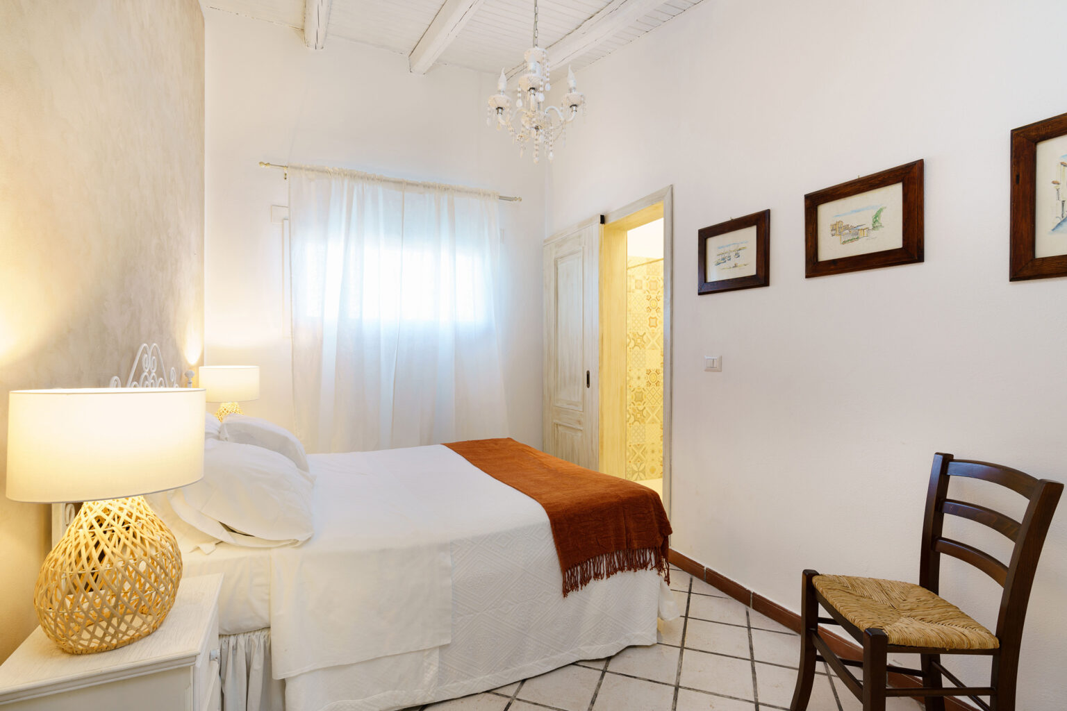 Cozy bedroom featuring a white bed with an orange throw, a wooden chair, decorative wall art, and a bedside lamp, illuminated by natural light from a window. | Only on SopranoVillas