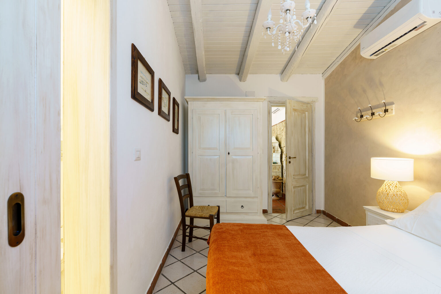 Cozy bedroom interior featuring a bed with an orange throw, a wooden chair, and a stylish light fixture. The room has white walls, decorative frames, and a wardrobe, creating a warm and inviting atmosphere. | Only on SopranoVillas