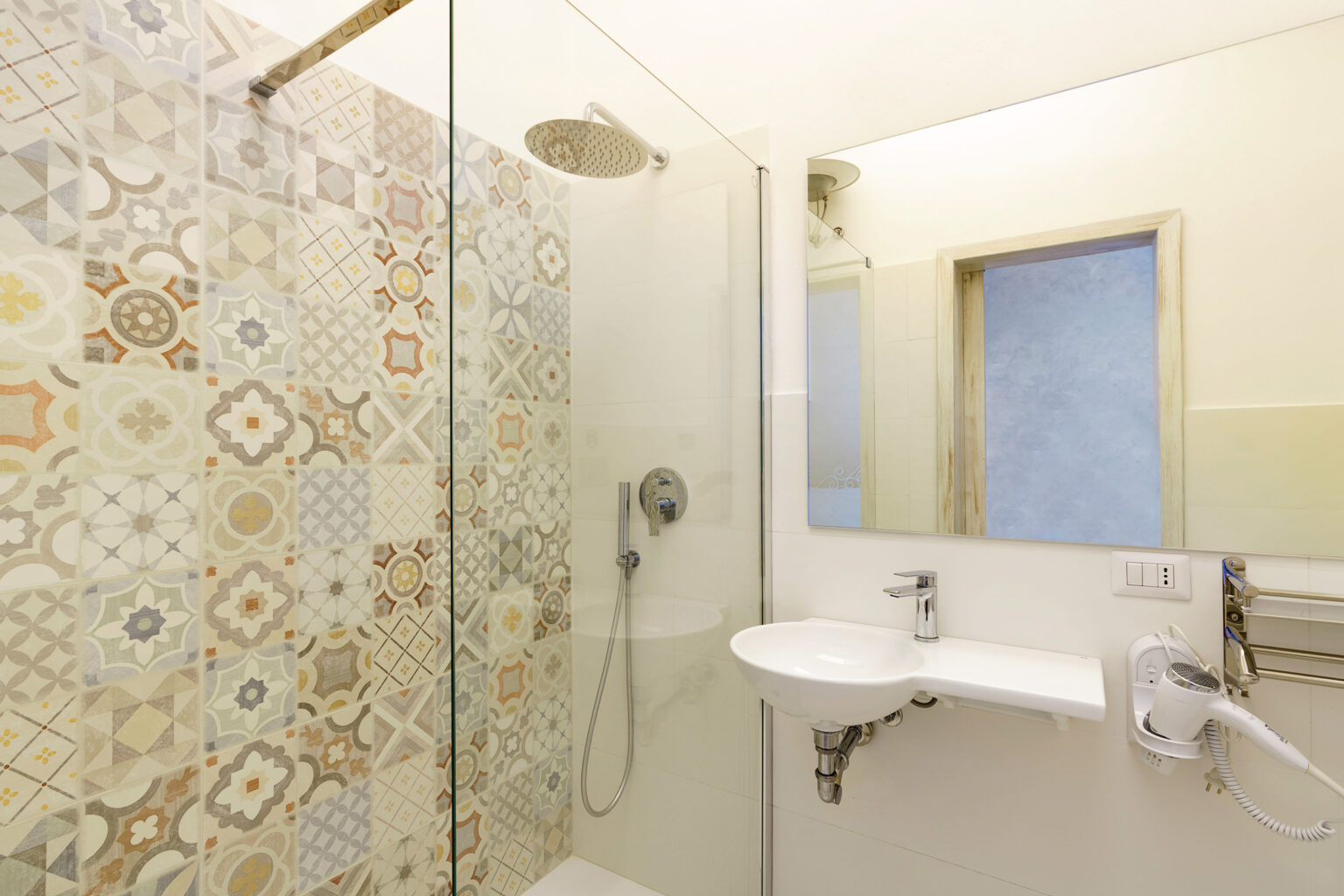 Modern bathroom featuring a walk-in shower with decorative tiled walls, a sleek sink, and a large mirror. Natural light streams in through a window, enhancing the contemporary design. | Only on SopranoVillas