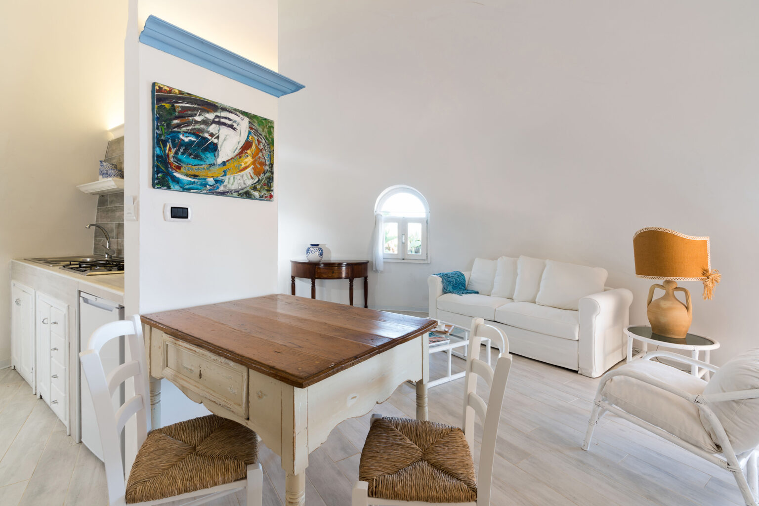 Cozy and modern living space featuring a wooden dining table with white chairs, a comfortable white sofa, and a colorful wall art piece. The room has bright, natural light and a minimalist design, ideal for relaxation. | Only on SopranoVillas