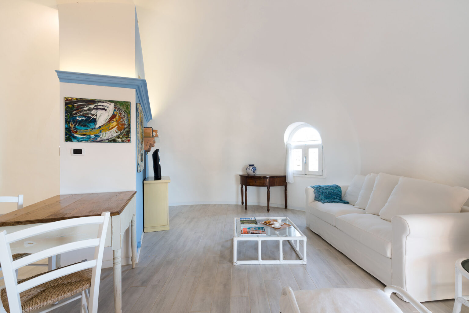 Bright and airy living room featuring a white sofa, wooden dining table, and an artistic wall decor. Natural light pours in through a unique arched window, enhancing the warm, inviting atmosphere. Ideal for relaxation and leisure. | Only on SopranoVillas