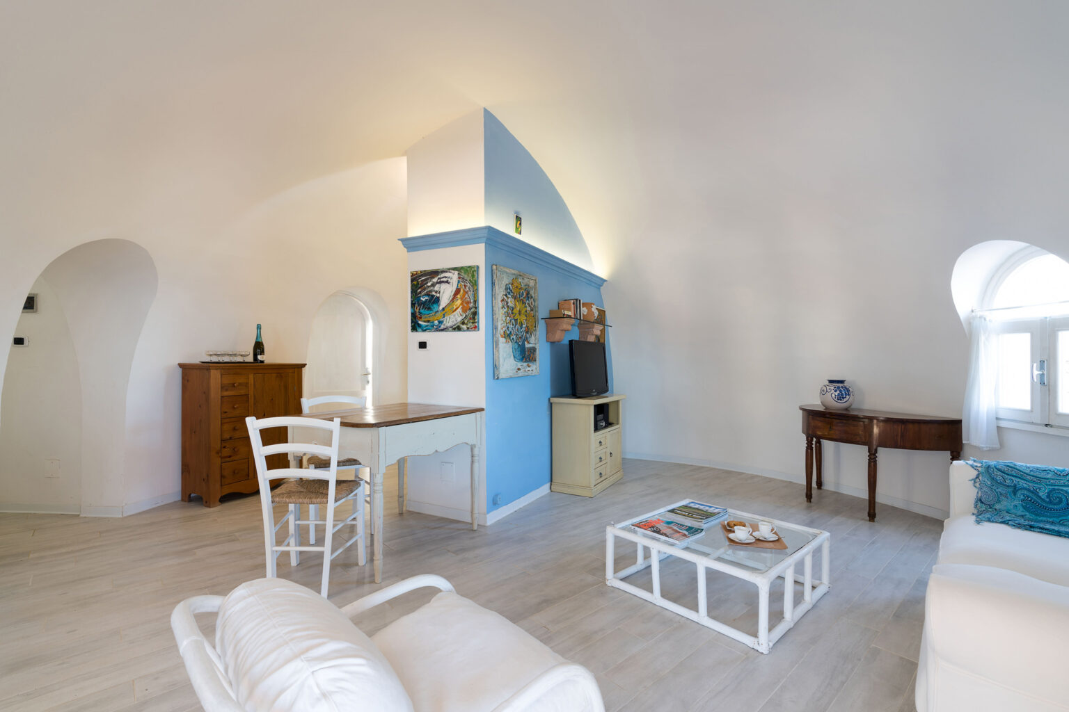 Bright and airy living room featuring white furniture, a light wood floor, and colorful accent walls. The space includes a dining table, a television, and decorative artwork, creating a cozy and inviting atmosphere ideal for relaxation. | Only on SopranoVillas