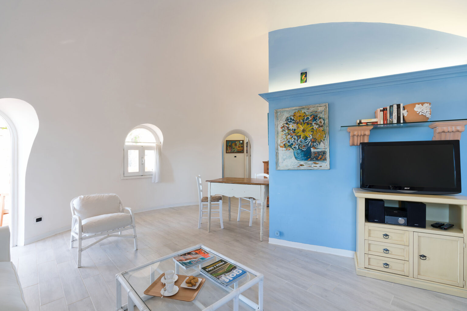 Bright and airy living room featuring a blue accent wall, modern furniture, and a television. The space includes a dining area with a table and chairs, decorative art pieces, and large windows allowing natural light. Ideal for a cozy and stylish atmosphere. | Only on SopranoVillas