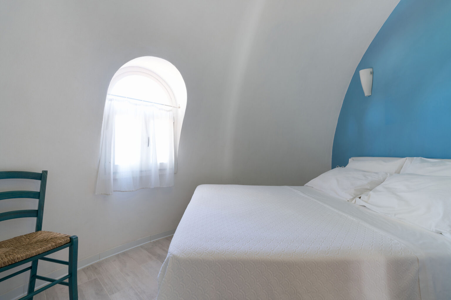 Cozy bedroom featuring a white bed, blue accent wall, and a traditional green chair, with natural light filtering through a small arched window. Ideal for a relaxing stay. | Only on SopranoVillas