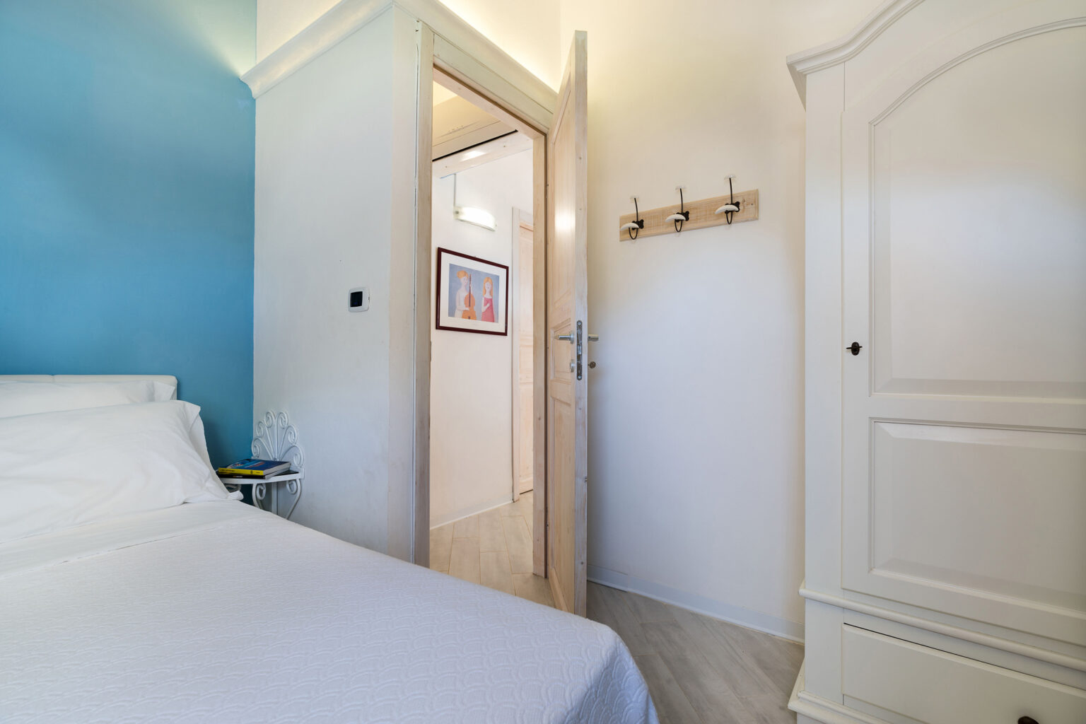 Cozy bedroom featuring a white bed, blue accent wall, and a doorway leading to a hallway. The room includes a white wardrobe and a wall-mounted coat rack, creating a welcoming atmosphere. | Only on SopranoVillas