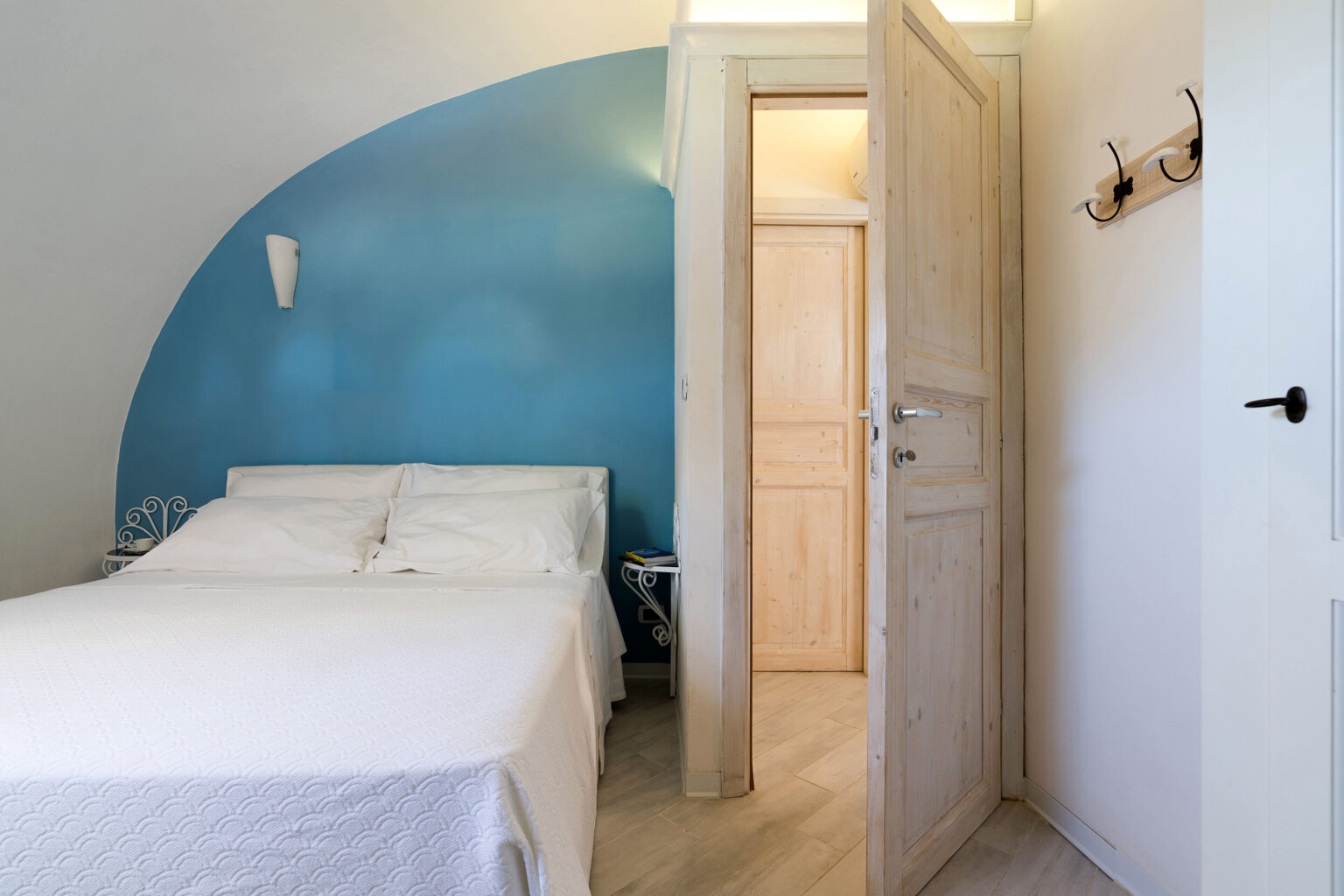 Cozy bedroom featuring a white bedspread, blue accent wall, and wooden door. The room includes a bedside table with decorative details and an open doorway leading to a wooden closet. Ideal for relaxation and comfort. | Only on SopranoVillas