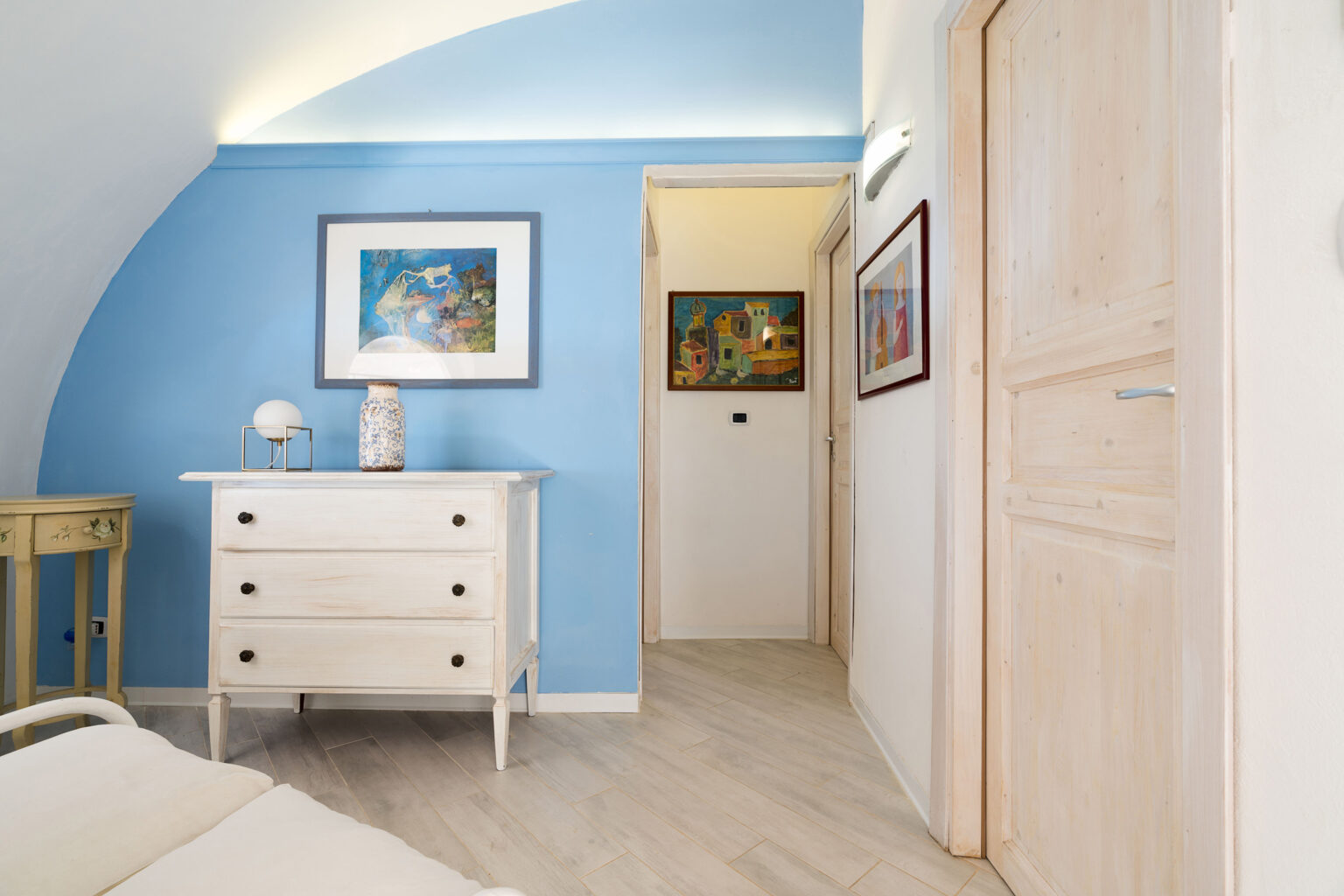 Cozy interior of a modern bedroom featuring a blue accent wall, a white dresser with decorative items, and artwork on the walls. The room has a warm ambiance with natural light and a stylish design, showcasing wooden flooring and an arching ceiling. | Only on SopranoVillas