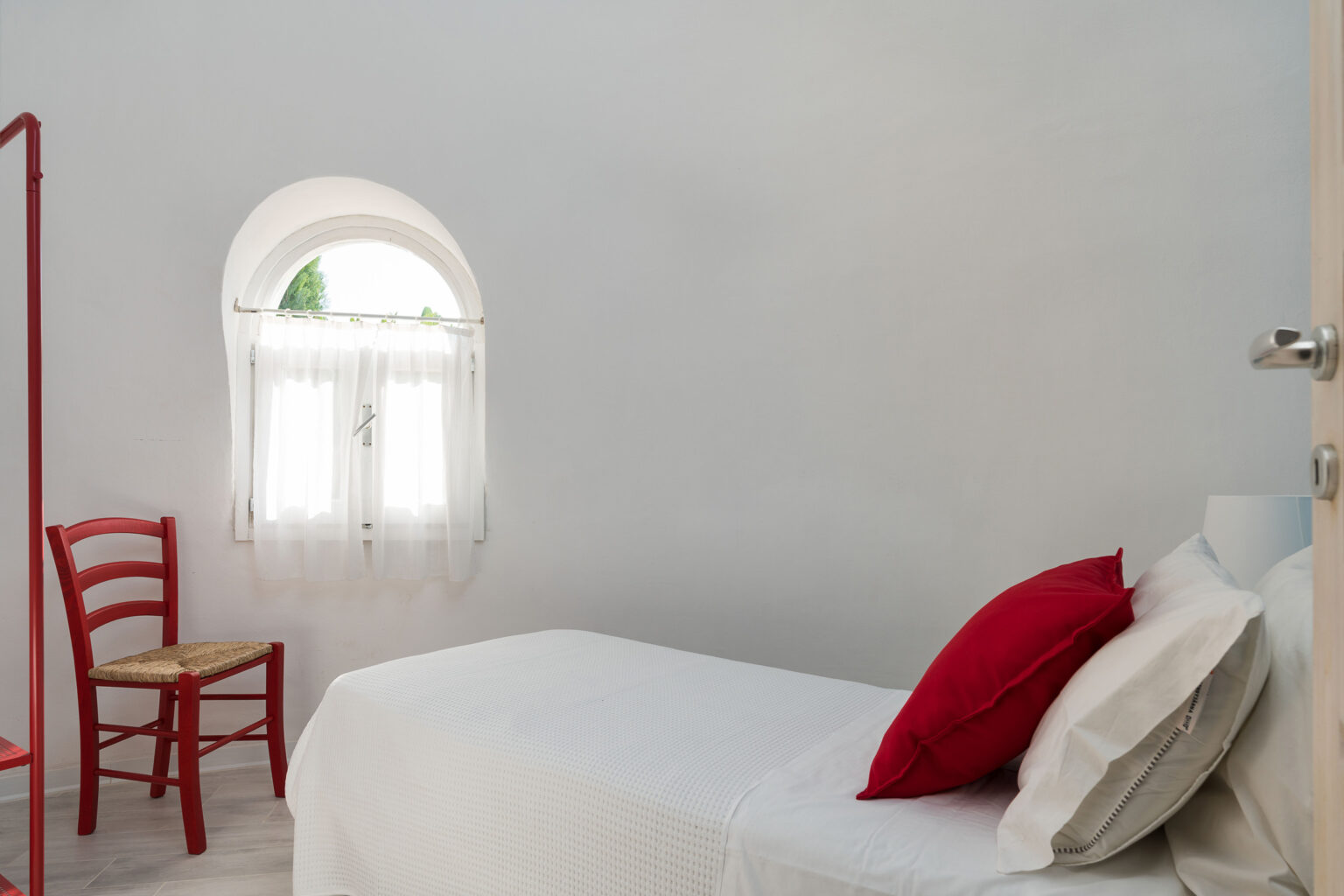 Minimalist bedroom featuring a single bed with white bedding and red decorative pillows, a red wooden chair, and a bright window adorned with sheer curtains, all set against a clean white wall. | Only on SopranoVillas
