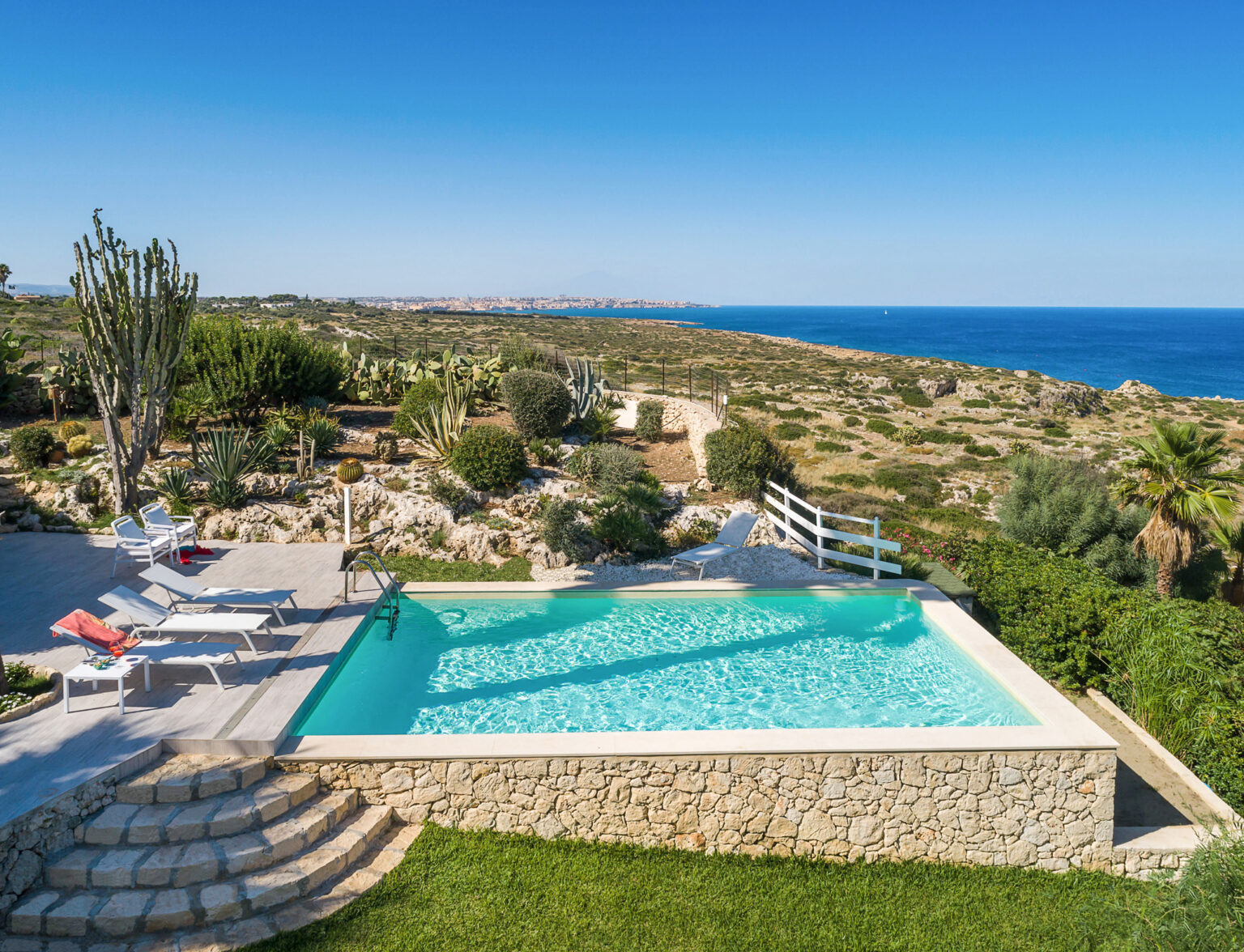 Stunning view of a luxury villa with an infinity pool overlooking the ocean, surrounded by lush greenery and rocky terrain. Ideal for vacation rentals or getaway retreats. | Only on SopranoVillas