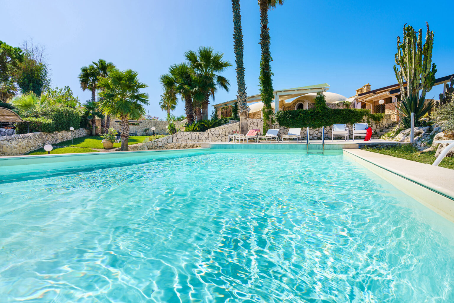 Crystal-clear swimming pool surrounded by palm trees and lush greenery, with a sunny blue sky and a relaxing atmosphere, ideal for leisure and vacation. | Only on SopranoVillas