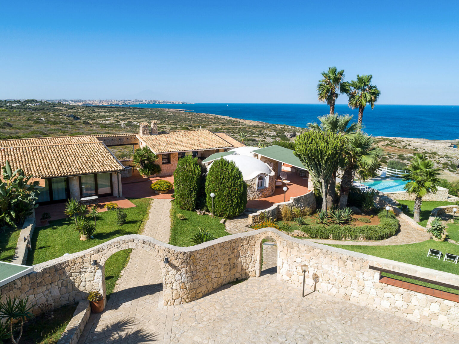 Aerial view of a luxurious coastal villa featuring a landscaped garden, palm trees, and a swimming pool, overlooking the blue ocean and scenic coastline. | Only on SopranoVillas