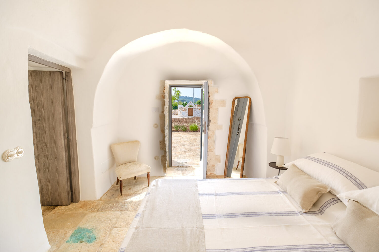 Bright, airy bedroom with white walls, a cozy bed, and a mirror, featuring an open door leading to a scenic outdoor view. Ideal for vacation rentals or interior design inspiration. | Only on SopranoVillas