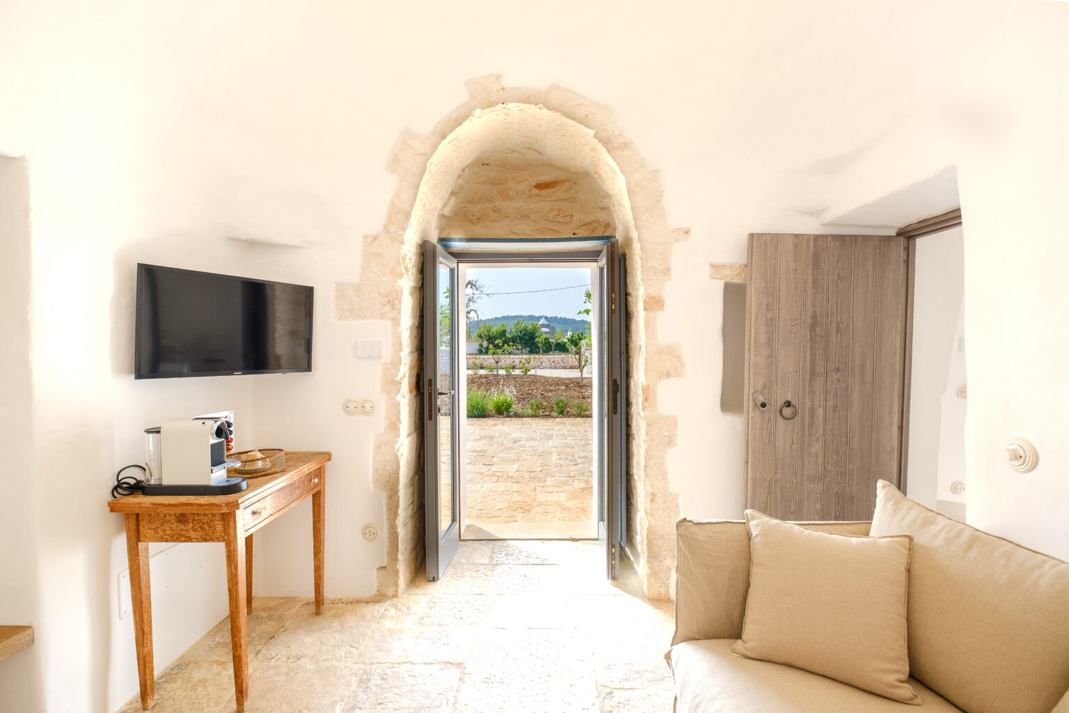 Interior view of a cozy room featuring a stone archway leading to an outdoor landscape, a wooden table with a coffee maker, a television mounted on the wall, and a comfortable beige sofa, all designed with a bright and airy ambiance. | Only on SopranoVillas