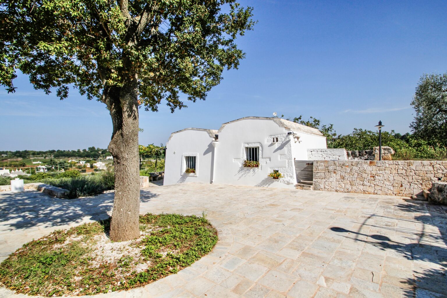 Charming whitewashed villa surrounded by lush greenery and a stone pathway, set against a clear blue sky in a tranquil landscape. | Only on SopranoVillas