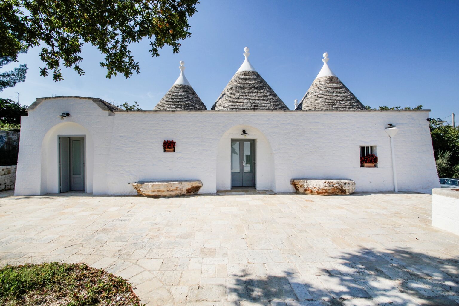 Trullo house with conical roofs in a sunny outdoor setting, featuring whitewashed walls and a stone patio, surrounded by greenery. | Only on SopranoVillas