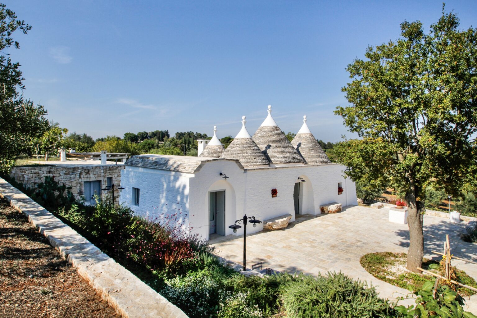 Traditional trulli house in Alberobello, Italy, featuring iconic conical roofs surrounded by lush greenery and a clear blue sky. | Only on SopranoVillas