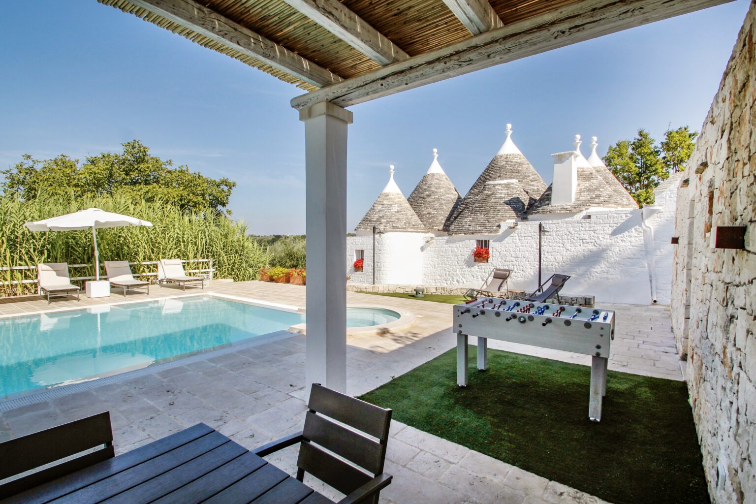 Scenic outdoor area featuring a swimming pool, lounge chairs, and a foosball table, with traditional trullo houses in the background surrounded by greenery. Ideal for relaxation and leisure activities. | Only on SopranoVillas