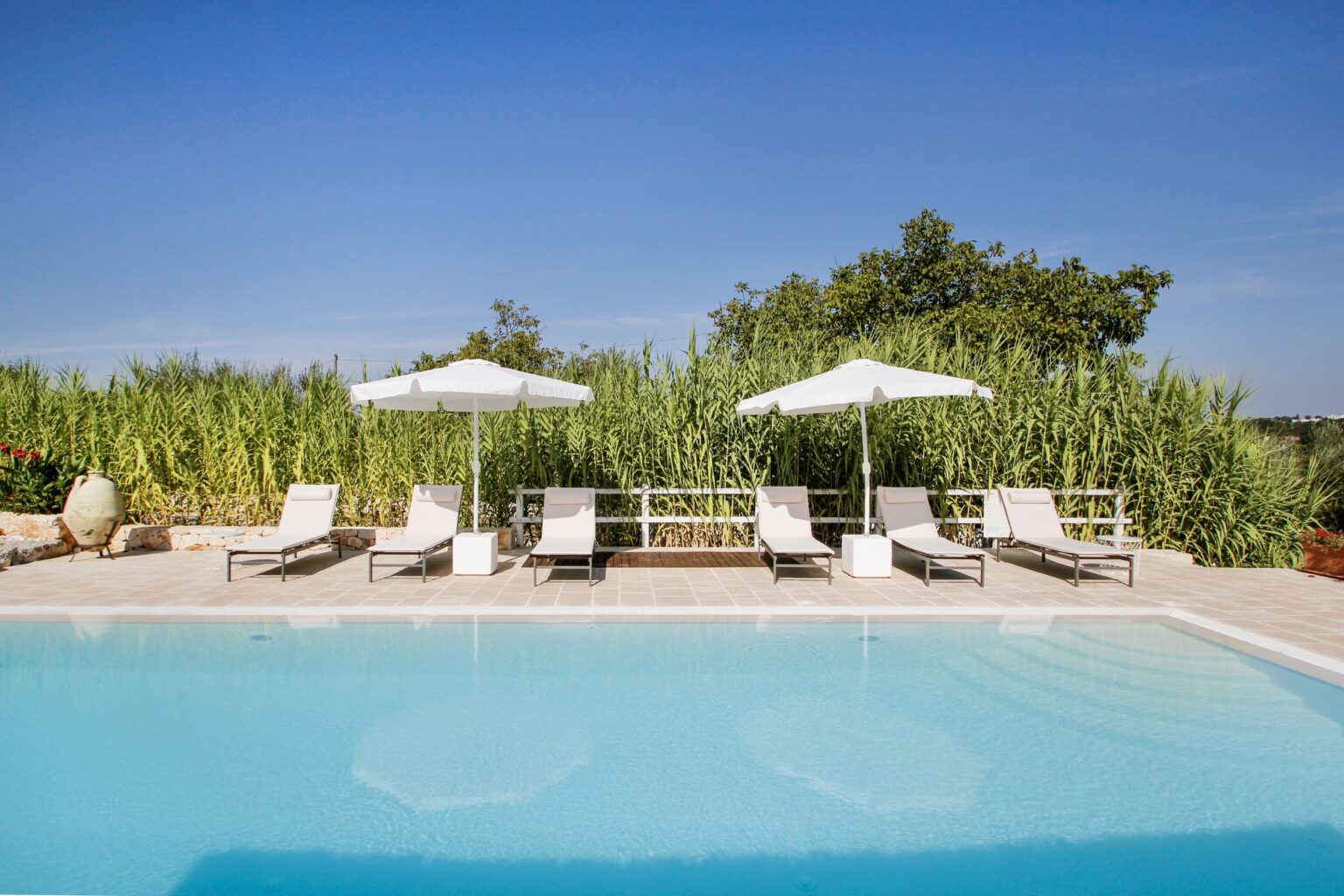 Serene outdoor swimming pool area featuring modern lounge chairs and white umbrellas, surrounded by lush greenery and a clear blue sky. Ideal for relaxation and leisure. | Only on SopranoVillas