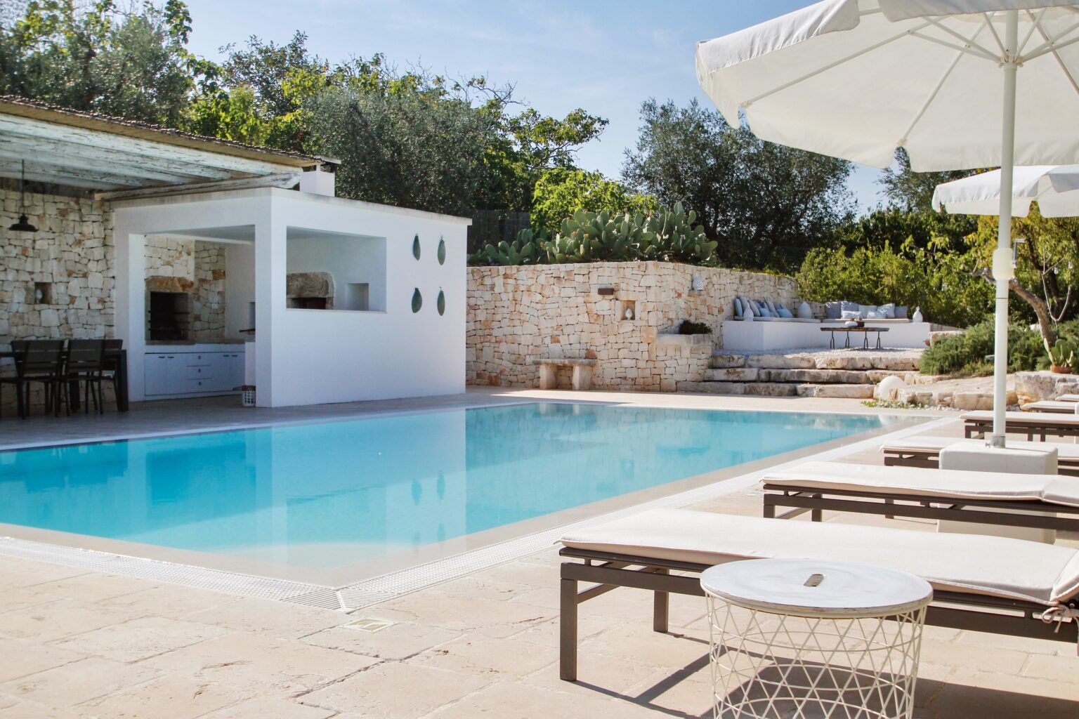 Modern outdoor swimming pool surrounded by lounge chairs and umbrellas, with a stone wall and landscaped greenery in the background, ideal for relaxation and leisure activities. | Only on SopranoVillas