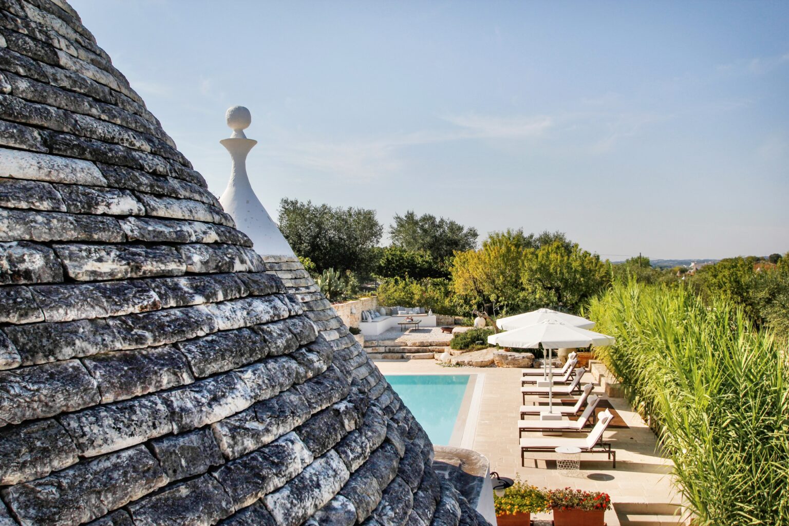 View of a traditional trullo roof with a swimming pool and sun loungers in a scenic garden setting, showcasing lush greenery and a clear sky. Ideal for vacation rentals in Puglia, Italy. | Only on SopranoVillas