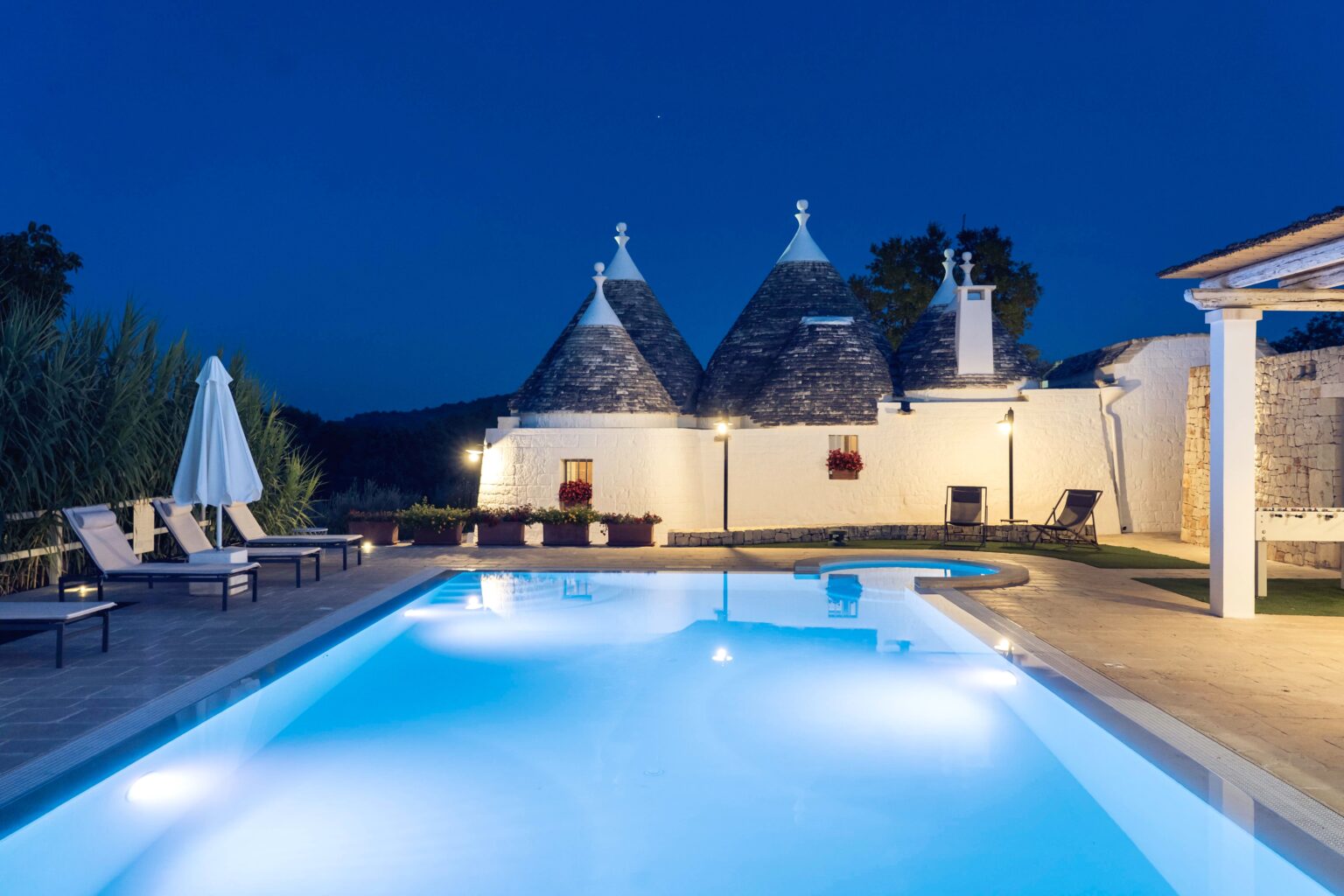 Tranquil nighttime view of a luxury villa featuring traditional trullo architecture, illuminated swimming pool, and comfortable lounge chairs, surrounded by lush greenery. | Only on SopranoVillas