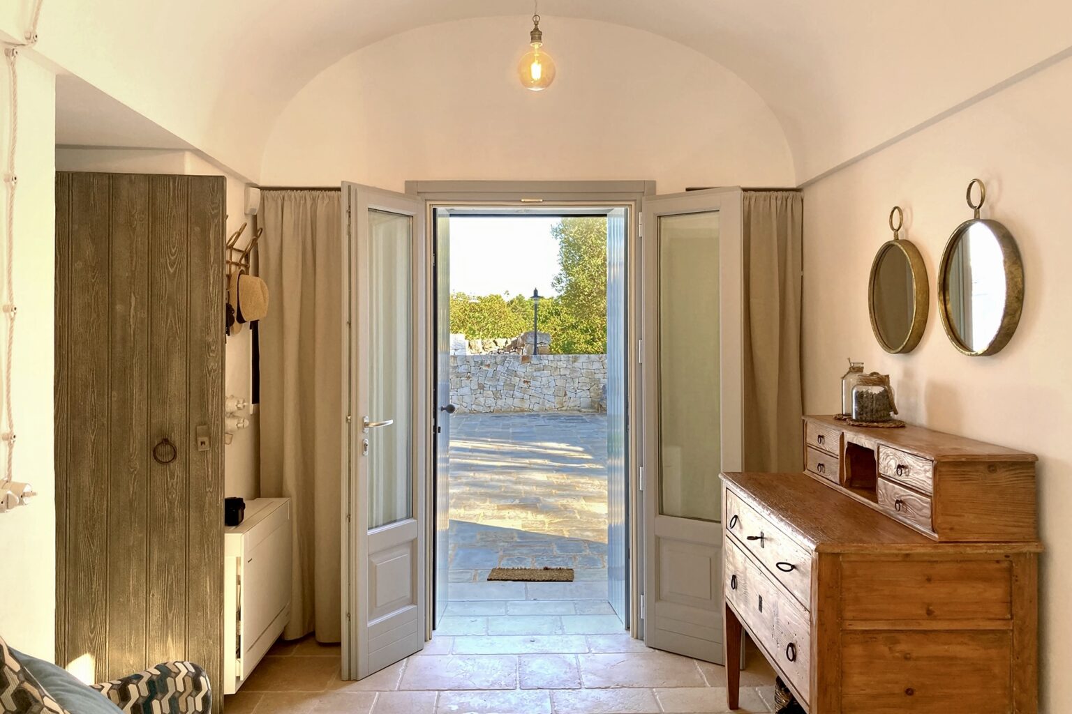Bright and inviting interior view of a room with open double doors leading to an outdoor space, featuring a wooden dresser and decorative mirrors on the wall, highlighting a blend of natural light and rustic decor. | Only on SopranoVillas