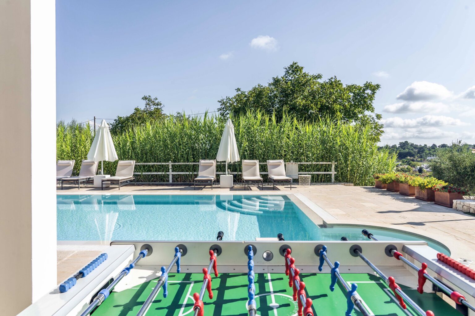 Outdoor pool area with lounge chairs and umbrellas, featuring a foosball table in the foreground, surrounded by lush greenery and a clear blue sky. Ideal for relaxation and recreation. | Only on SopranoVillas