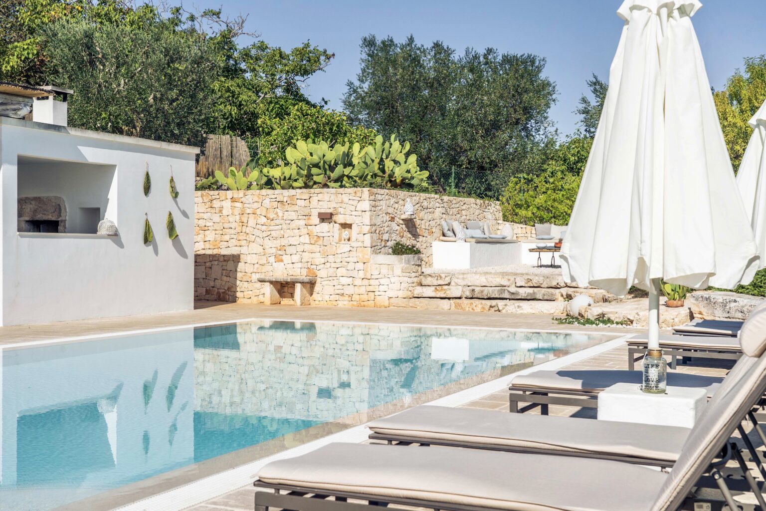 Serene outdoor pool area with lounge chairs and white umbrellas surrounded by lush greenery and stone walls, ideal for relaxation and leisure. | Only on SopranoVillas