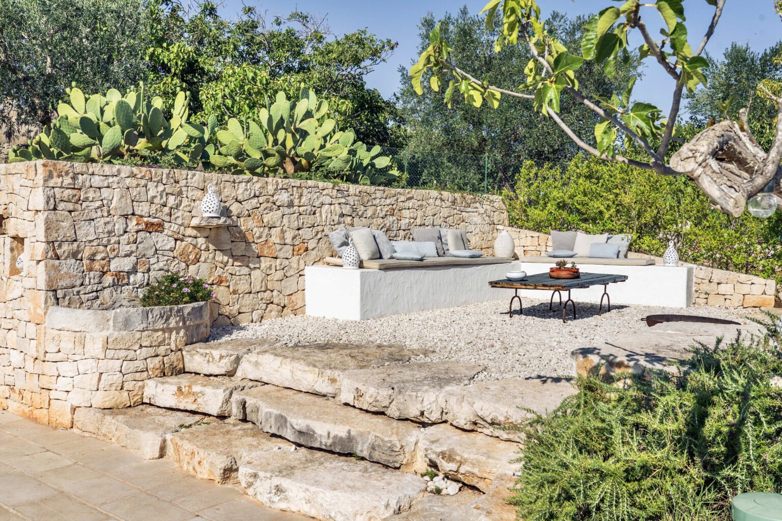 Outdoor living space featuring a stone wall, comfortable seating area with cushions, and a coffee table surrounded by lush greenery and cacti. Ideal for relaxation and entertaining in a natural setting. | Only on SopranoVillas