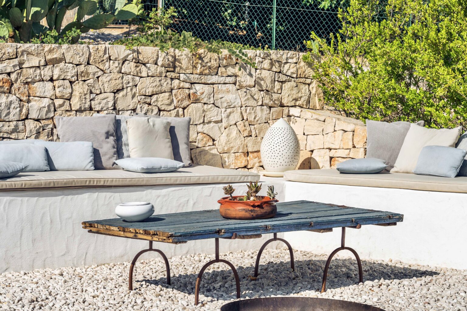 Outdoor lounge area featuring a stone wall, comfortable gray cushions on a white bench, a rustic wooden table with a clay pot of succulents, and decorative accents in a gravel setting. | Only on SopranoVillas