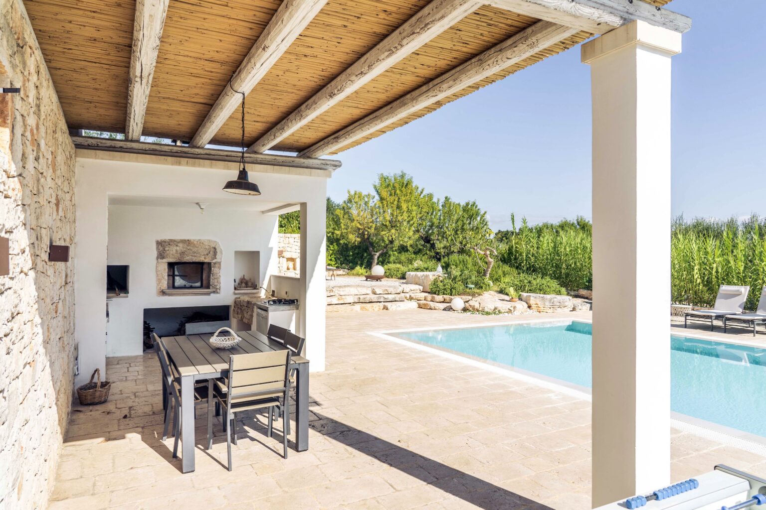 Outdoor dining area with a stone grill, surrounded by greenery, adjacent to a swimming pool under a shaded pergola. Ideal for relaxation and entertaining in a sunny environment. | Only on SopranoVillas