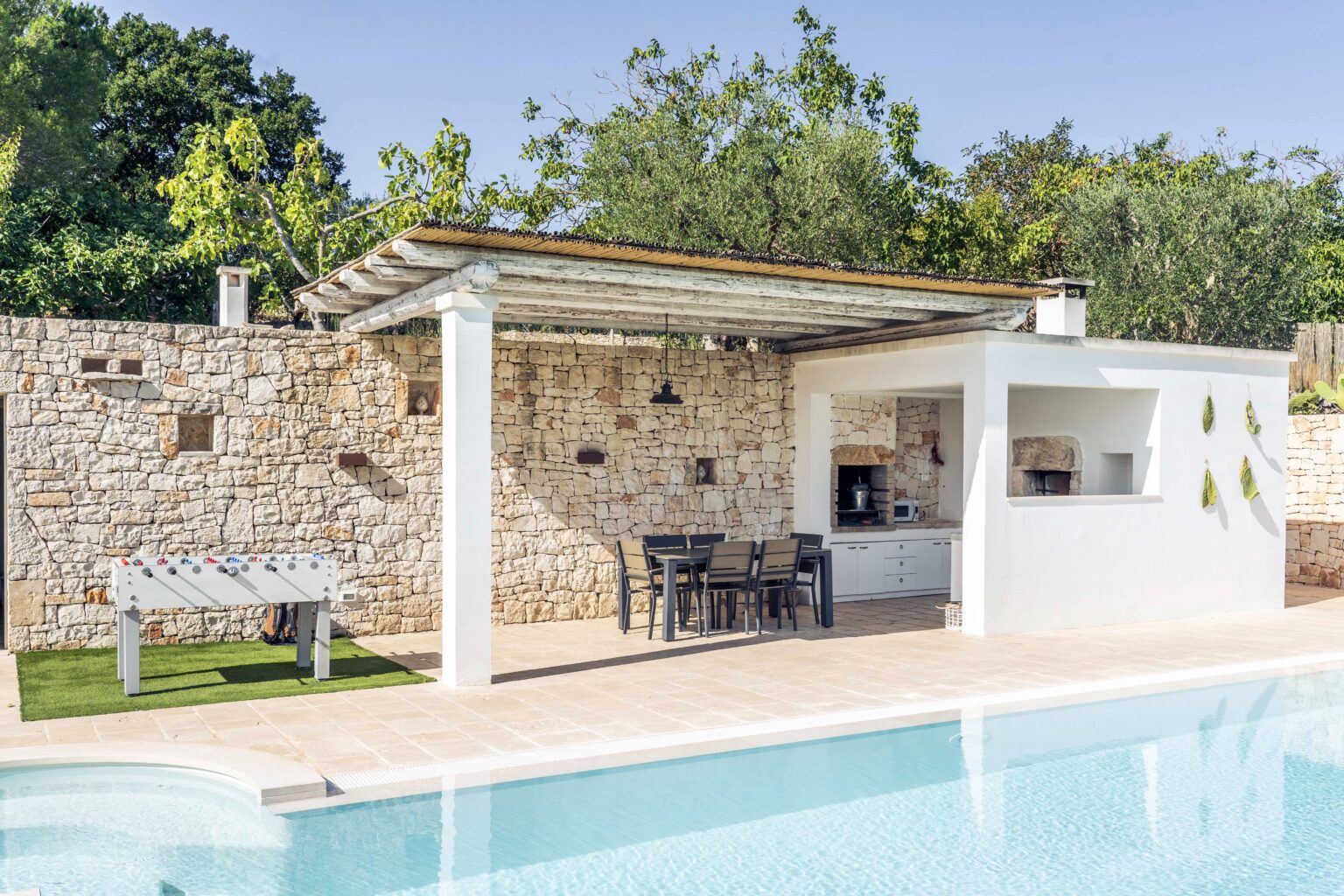 Modern outdoor pool area featuring a stone wall, shaded seating under a pergola, and a nearby outdoor kitchen, surrounded by lush greenery. Ideal for relaxation and entertaining. | Only on SopranoVillas