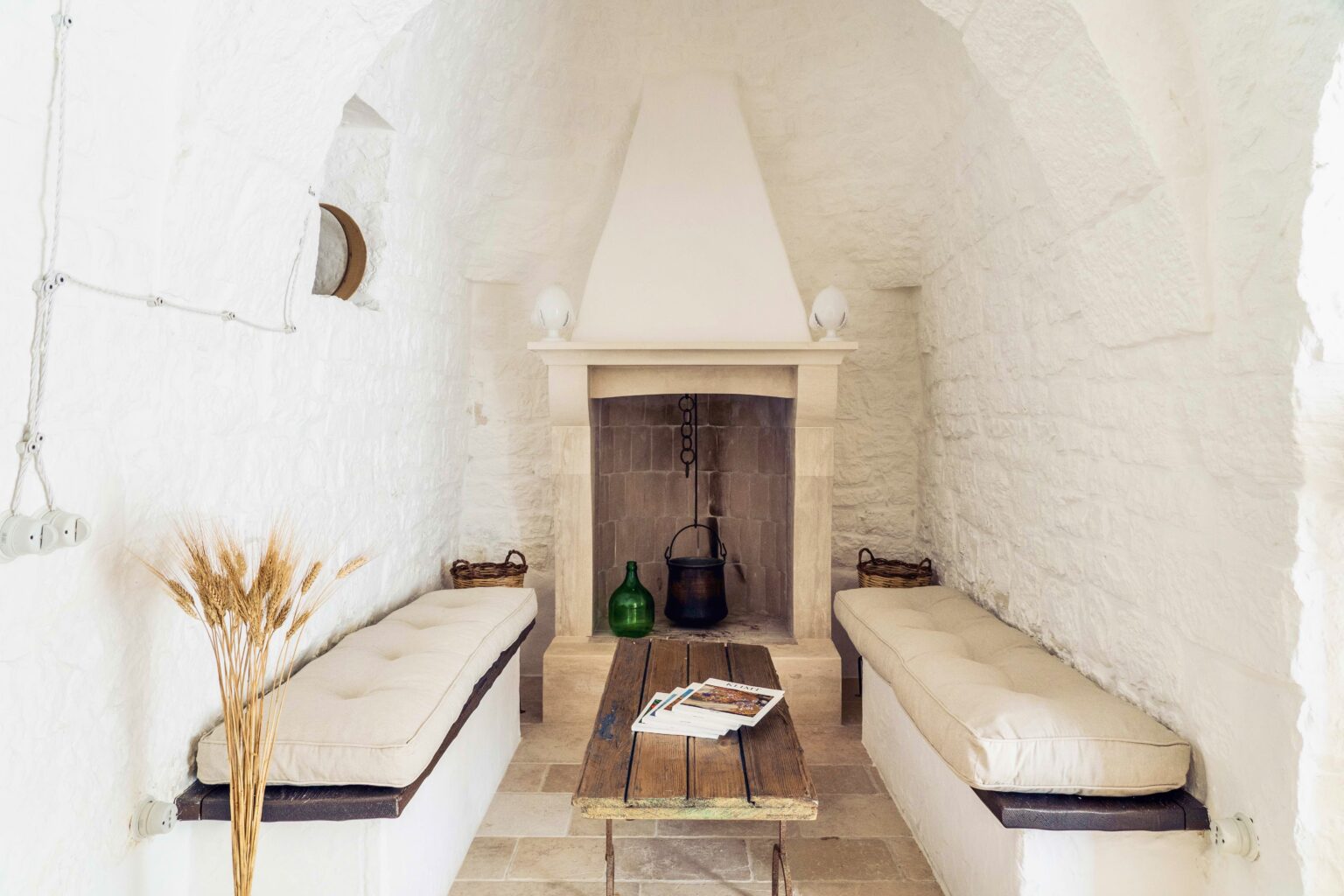 Cozy interior of a white stone room featuring a minimalist fireplace, two cushioned benches, and a rustic wooden table with magazines, enhancing a warm and inviting atmosphere. | Only on SopranoVillas