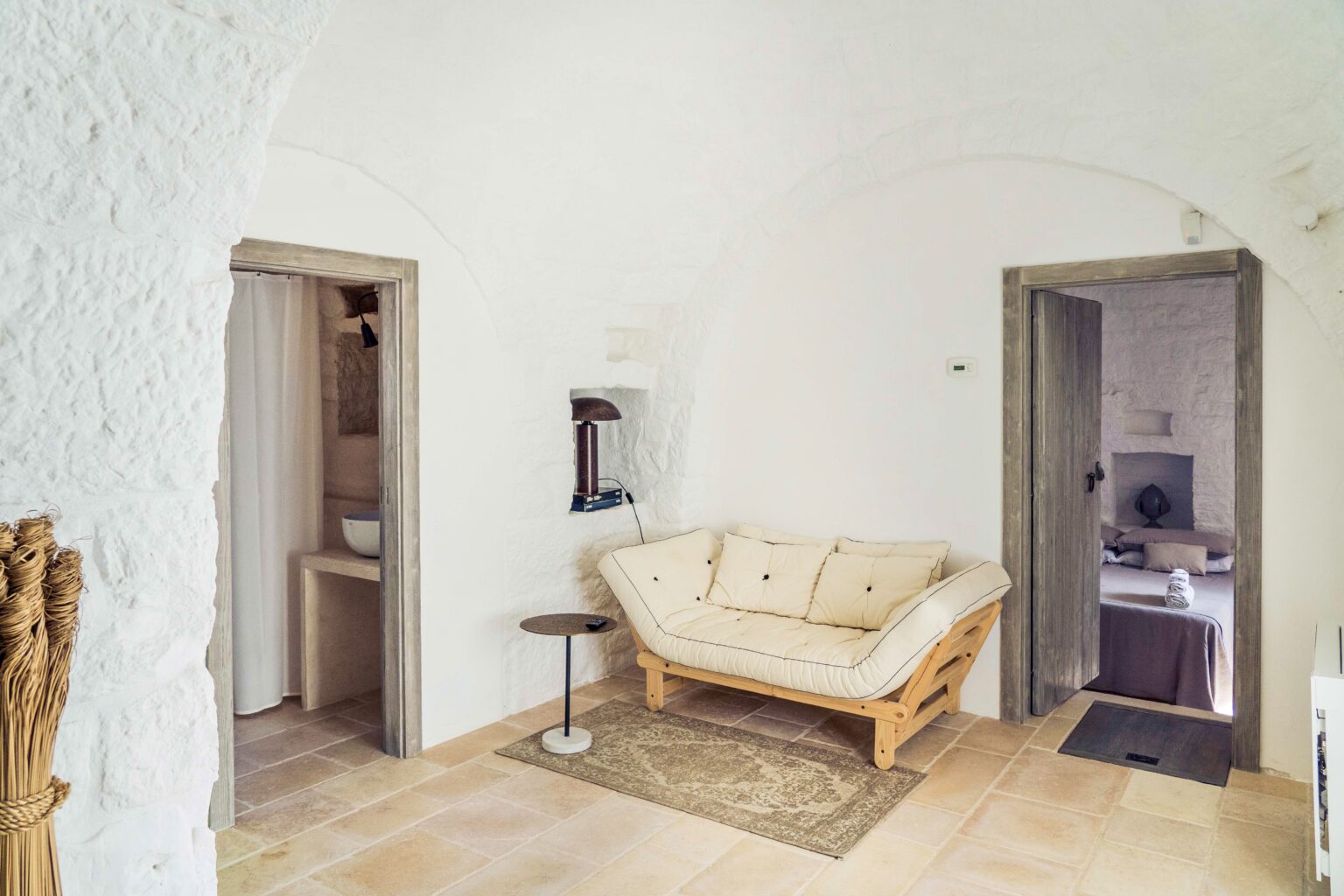 Cozy interior of a minimalist living space featuring a beige futon, a small side table, and stone walls. The room includes a doorway leading to a bathroom and another to a bedroom, showcasing a warm, inviting atmosphere with natural light. | Only on SopranoVillas