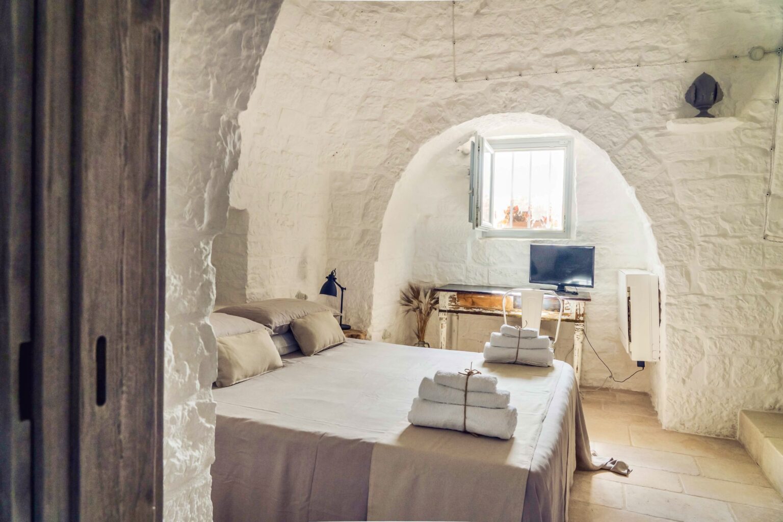 Cozy bedroom in a rustic stone cottage featuring a large bed with neatly arranged towels, a small workspace with a computer, and an arched ceiling that enhances the charming atmosphere. Natural light streams in through a window, creating a warm and inviting space. | Only on SopranoVillas