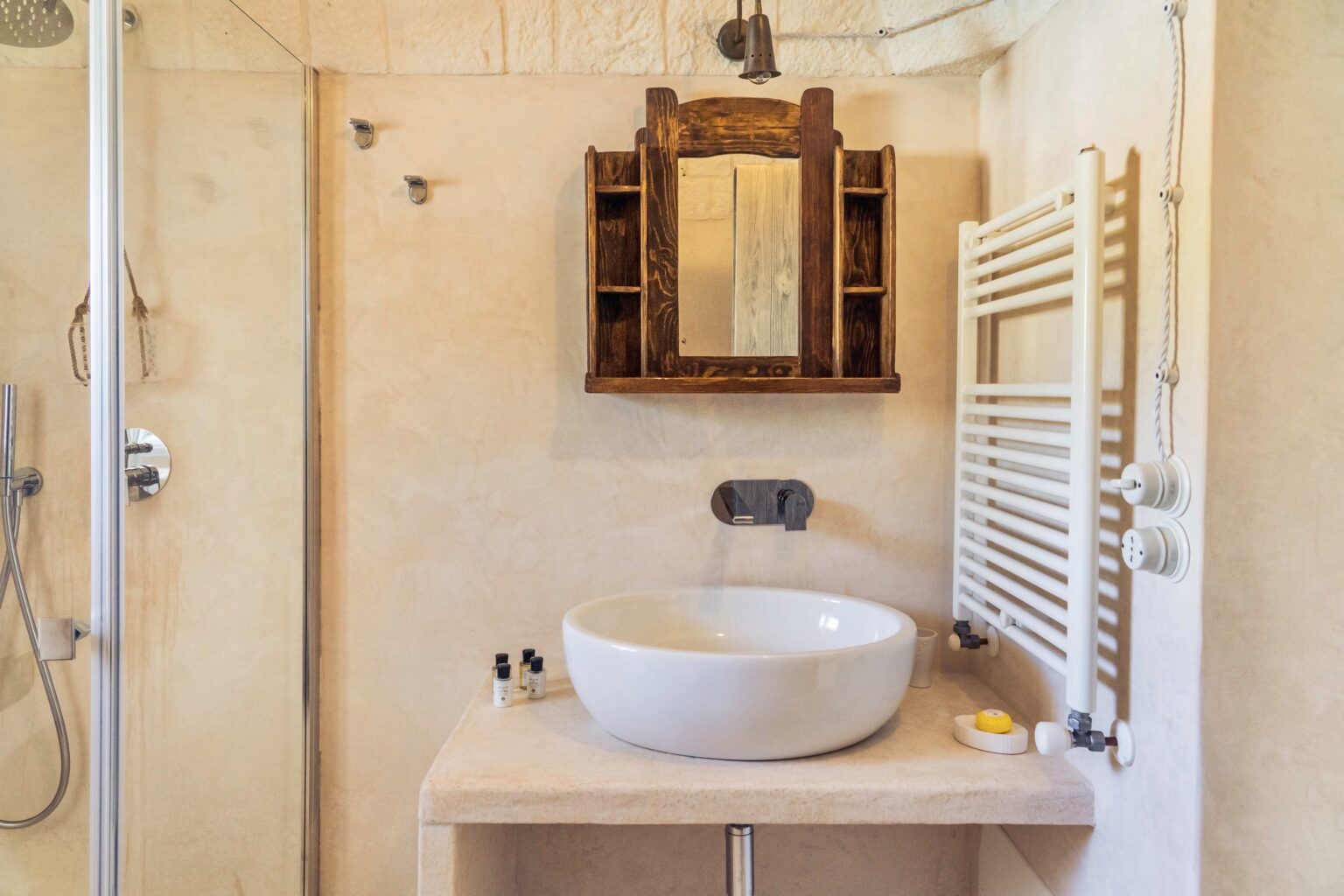 Modern bathroom interior featuring a stylish white vessel sink on a wooden countertop, a vintage mirror above, a towel radiator, and a glass shower enclosure, offering a blend of contemporary and rustic design elements. | Only on SopranoVillas