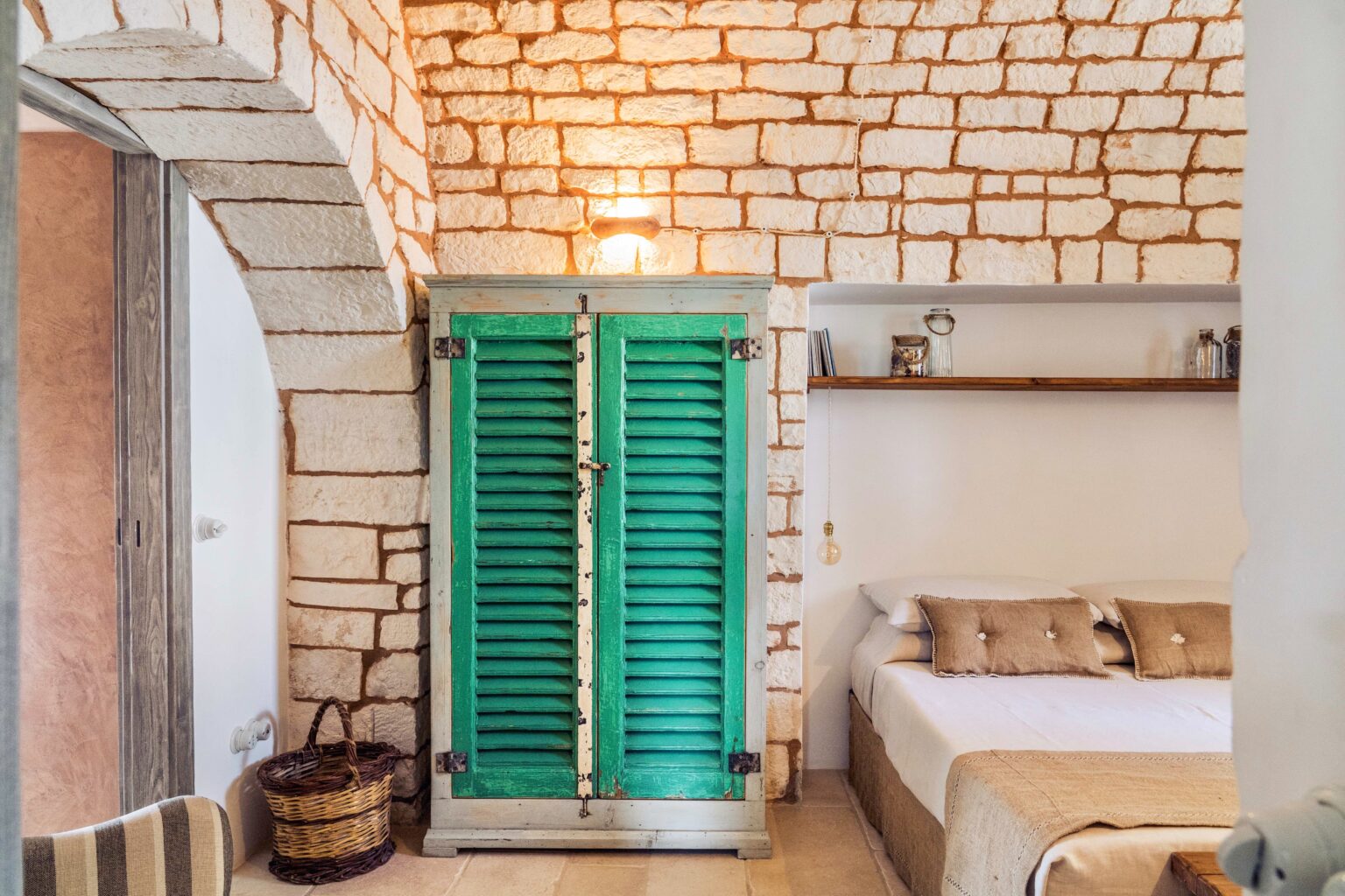 Interior view of a cozy bedroom featuring a stone wall, a green wooden wardrobe, and a minimalist bed with beige linens, creating a warm and inviting atmosphere. | Only on SopranoVillas