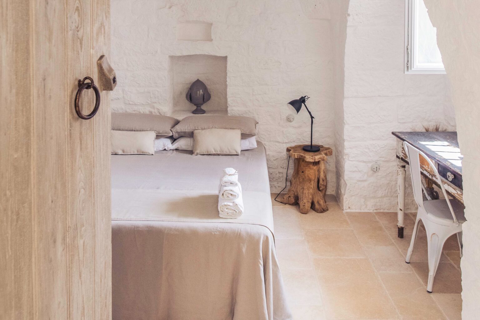 Cozy and modern bedroom interior featuring a neutral color palette, a comfortable bed with soft linens, a rustic wooden side table, and a stylish desk with a lamp, all set against whitewashed walls and warm tile flooring. Ideal for a relaxing getaway or home inspiration. | Only on SopranoVillas