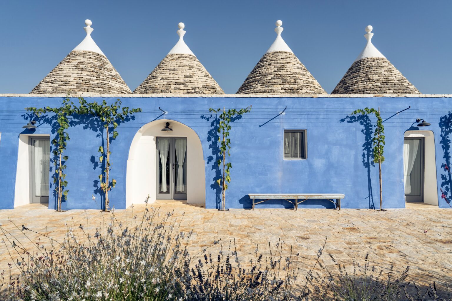 Charming blue trullo house with conical stone roofs, surrounded by lush greenery and blooming lavender, set against a clear blue sky. | Only on SopranoVillas