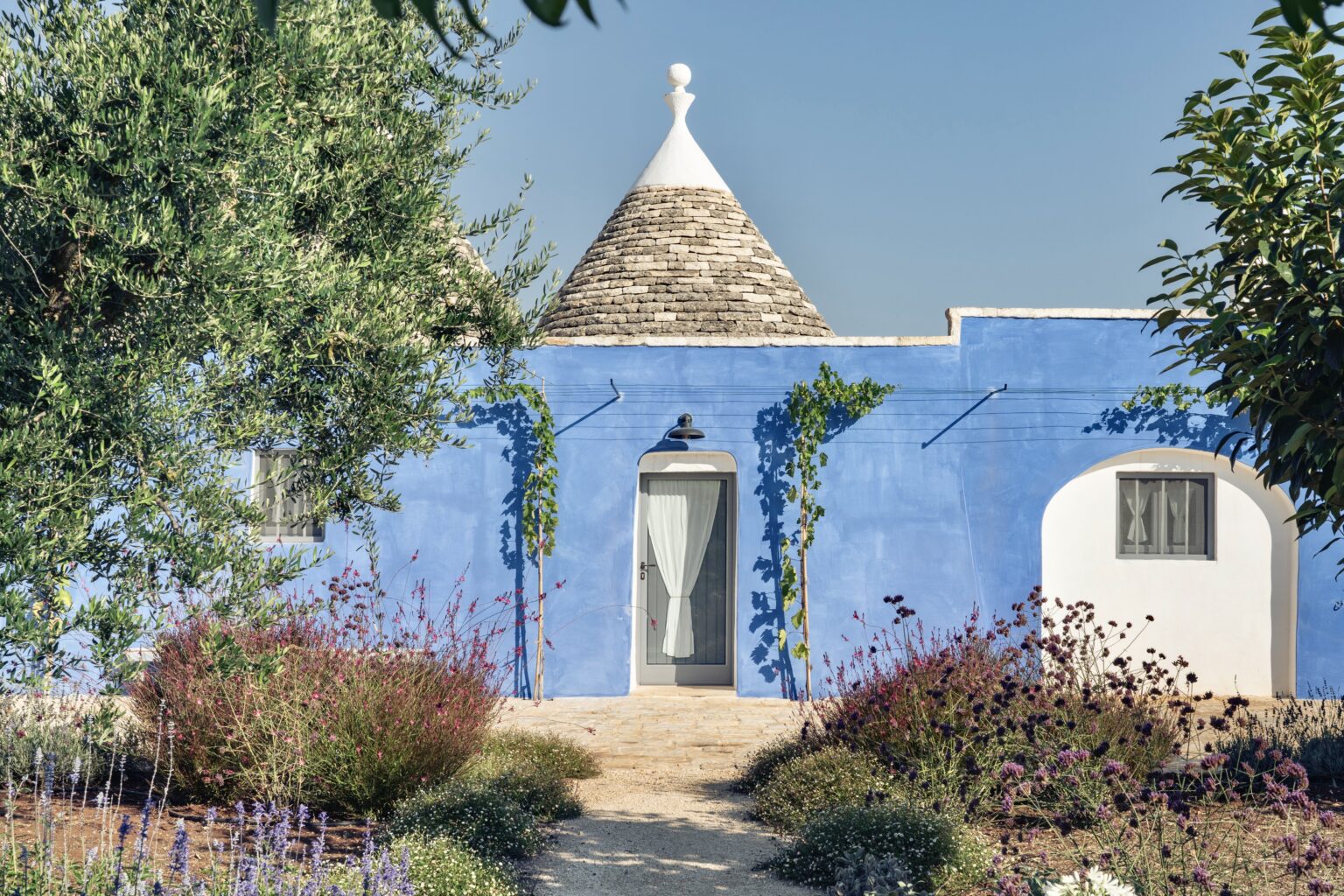 Charming blue trullo house surrounded by vibrant gardens in Puglia, Italy, featuring a traditional conical roof and lush greenery. | Only on SopranoVillas