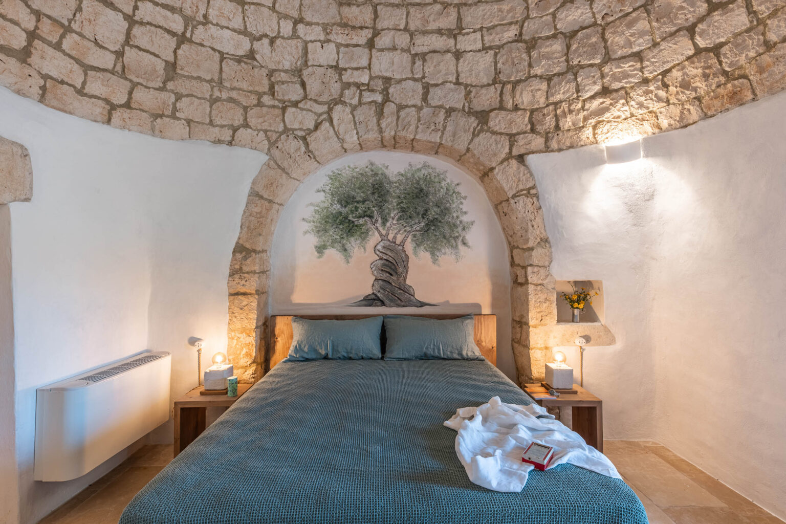 Cozy bedroom featuring a stone arch ceiling, a wooden bed with blue bedding, and a decorative mural of a twisted tree. The room includes bedside tables with lamps, a small floral arrangement, and a heater, creating a warm and inviting atmosphere. | Only on SopranoVillas