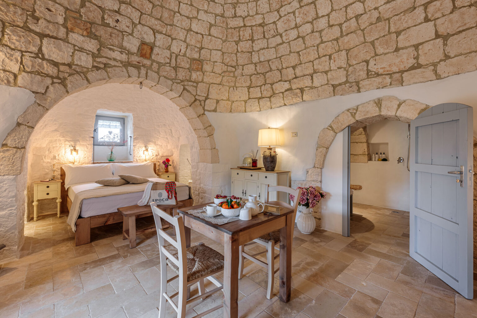 Cozy interior of a rustic stone bedroom featuring an arched ceiling, a comfortable bed with decorative pillows, a wooden bench, and a dining table set for breakfast with fresh fruit and tea. The room is illuminated by soft lighting and decorated with flowers, creating a warm and inviting atmosphere. | Only on SopranoVillas
