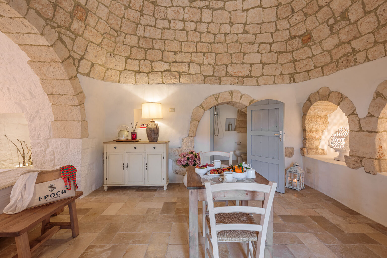 Interior view of a rustic dining area featuring a stone arch ceiling, wooden dining table with fresh fruits, white wooden chairs, and a light-colored sideboard, creating a warm and inviting atmosphere. | Only on SopranoVillas