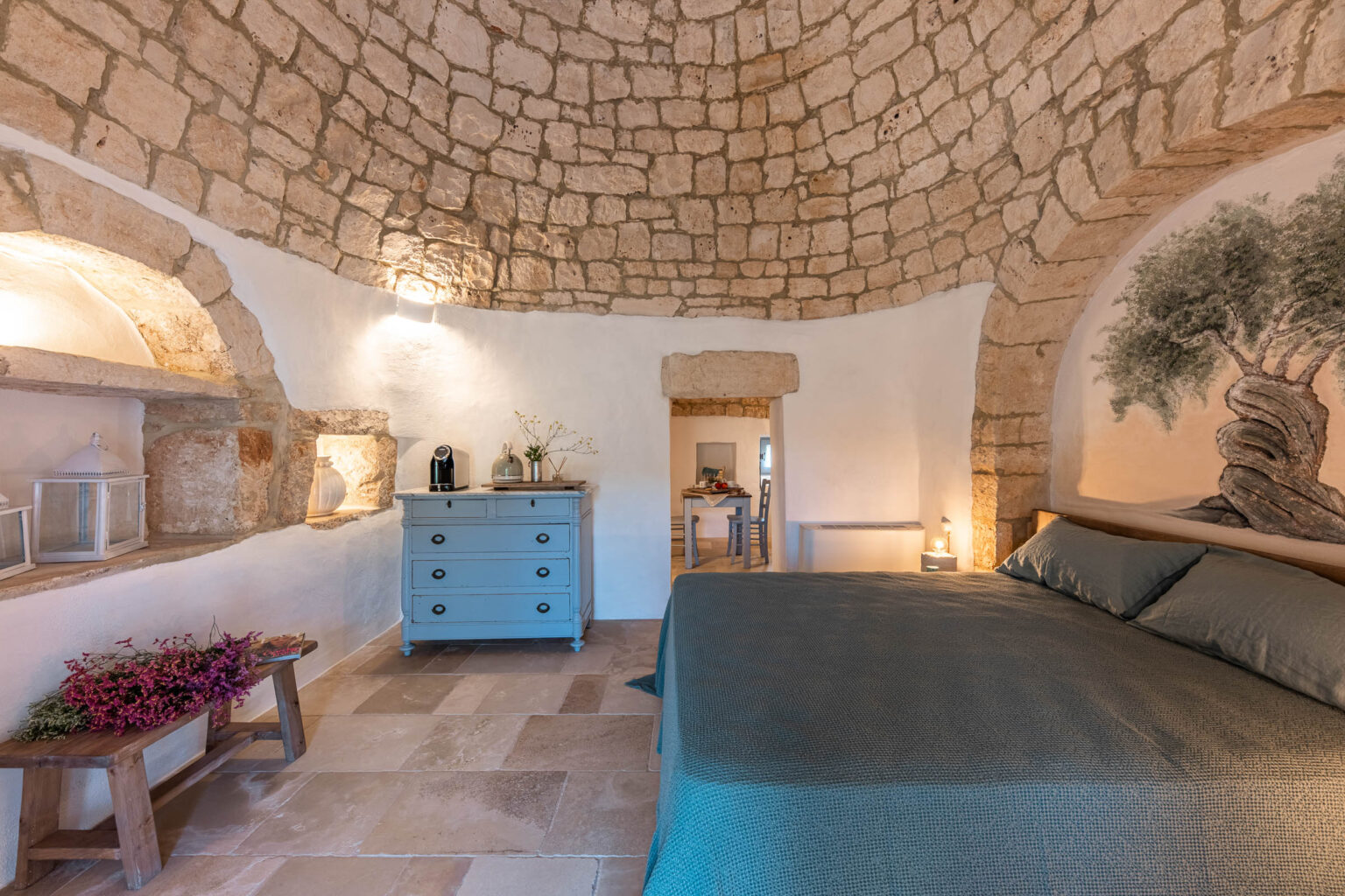 Interior of a cozy, uniquely designed bedroom featuring a curved stone ceiling, a light blue dresser, and a large bed with a textured blanket. The room includes rustic decor elements like a wooden bench with dried flowers and a wall mural of an olive tree, creating a warm and inviting atmosphere. | Only on SopranoVillas